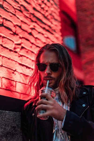 Beau jeune homme en lunettes de soleil et veste en cuir boire emporter cocktail de tasse en plastique sous la lumière rouge sur la rue — Photo de stock