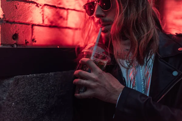 Attrayant jeune homme en lunettes de soleil et veste en cuir boire emporter cocktail de tasse en plastique sous la lumière rouge sur la rue — Photo de stock