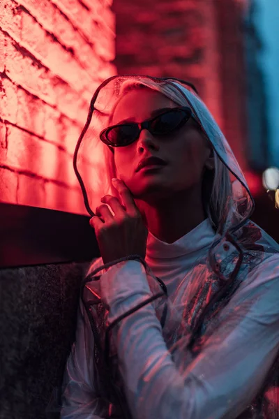 Belle jeune femme en imperméable transparent et lunettes de soleil dans la rue la nuit sous la lumière rouge — Photo de stock