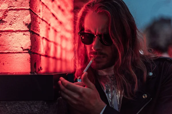 Joven guapo en gafas de sol y chaqueta de cuero fumar cigarrillo bajo la luz roja en la calle — Stock Photo