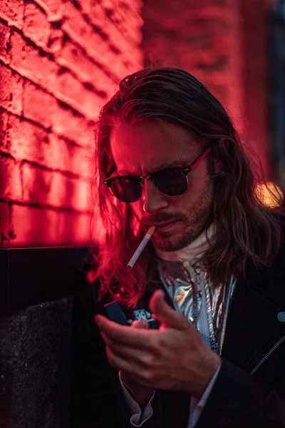 Attractive young man in sunglasses and leather jacket smoking cigarette under red light on street — Stock Photo