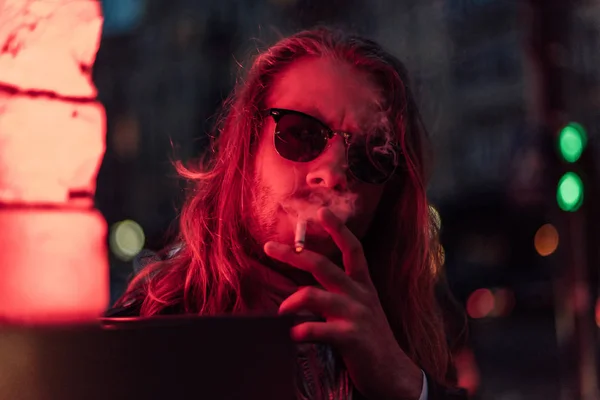 Schöner junger Mann mit Sonnenbrille raucht Zigarette unter Rotlicht auf der Straße — Stockfoto