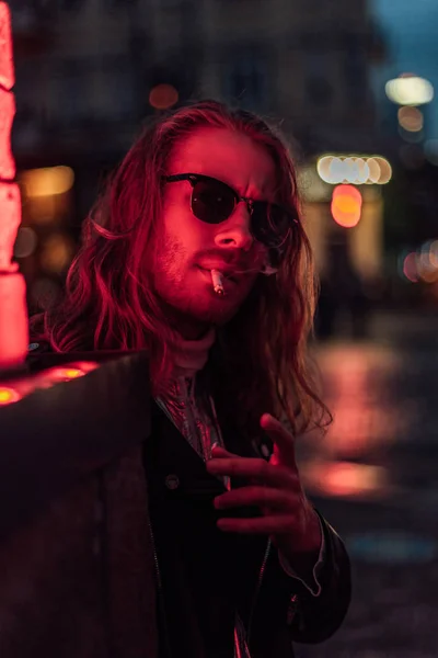 Joven con estilo en gafas de sol fumar cigarrillo bajo la luz roja en la calle - foto de stock