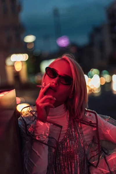 Красива молода жінка в прозорому плащі і сонцезахисних окулярах курить на вулиці вночі під червоним світлом — стокове фото