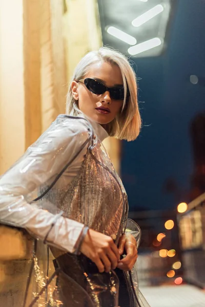 Красива молода жінка в прозорому плащі і сонцезахисних окулярах дивиться на камеру на вулиці вночі — стокове фото