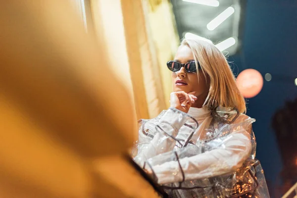 Hermosa joven en impermeable transparente y gafas de sol en la calle por la noche - foto de stock