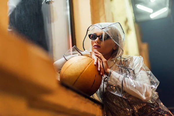 Bella giovane donna con pallone da basket in impermeabile trasparente e occhiali da sole in strada di notte — Foto stock