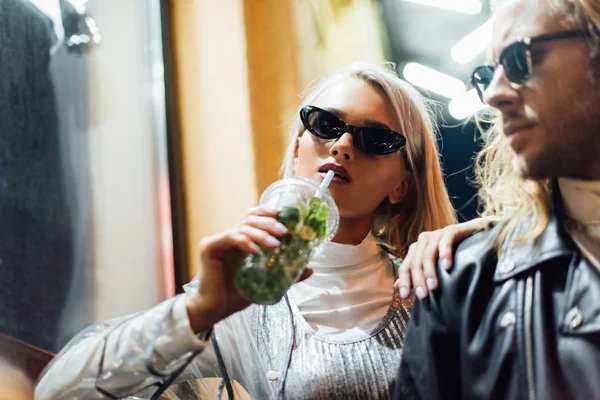 Giovane donna elegante in occhiali da sole bere mojito dalla tazza di plastica mentre trascorre del tempo con il fidanzato in strada di notte — Foto stock