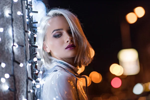 Bela jovem mulher inclinada na parede com guirlanda branca na rua da cidade à noite — Fotografia de Stock