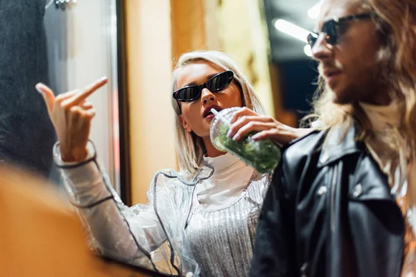 Jeune femme élégante dans les lunettes de soleil boire mojito de tasse en plastique et montrant majeur à la caméra tout en passant du temps avec petit ami dans la rue la nuit — Photo de stock