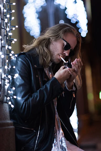 Atractivo joven en gafas de sol y chaqueta de cuero fumar cigarrillo bajo la guirnalda en la calle de la ciudad por la noche - foto de stock