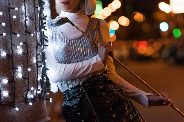 Tiro recortado de la mujer joven con estilo club de golf apoyado en la pared con guirnalda blanca en la calle de la ciudad por la noche - foto de stock