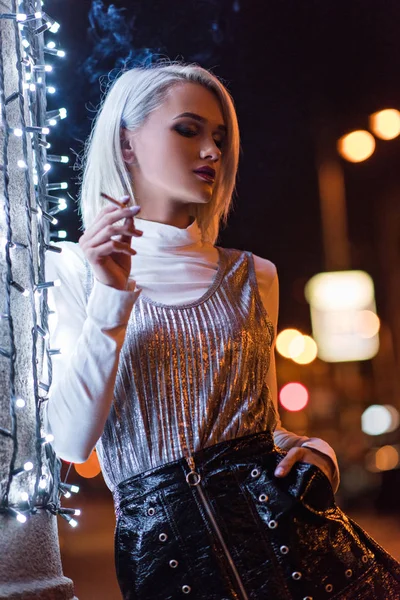 Hermosa joven mujer fumando en la calle por la noche mientras se apoya en la pared con guirnalda blanca - foto de stock