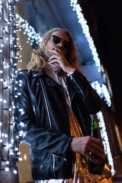 Bottom view of handsome young man in sunglasses and leather jacket holding bottle of beer and smoking cigarette under garland on street at night — Stock Photo