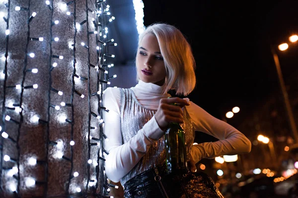 Young woman with bottle of beer on street at night leaning on wall with white garland — Stock Photo