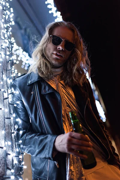 Bottom view of handsome young man in sunglasses and leather jacket holding bottle of beer and standing under garland on street at night — Stock Photo