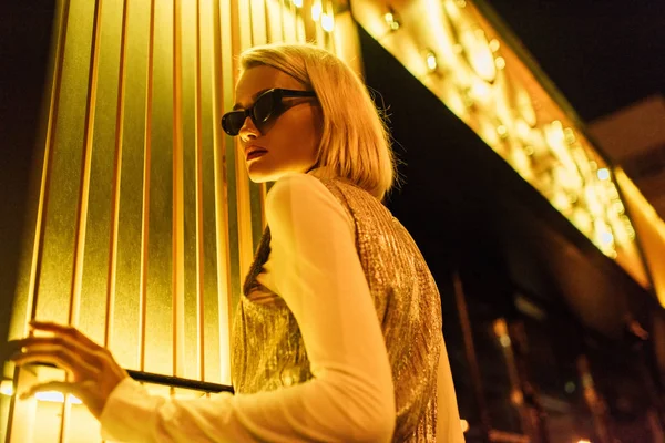 Bottom view of young woman in sunglasses and glossy top over turtleneck on street at night under yellow light — Stock Photo