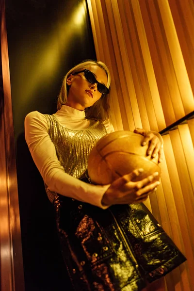 Visão inferior da jovem mulher elegante em óculos de sol com bola de basquete dourada brilhante na rua à noite sob luz amarela — Fotografia de Stock