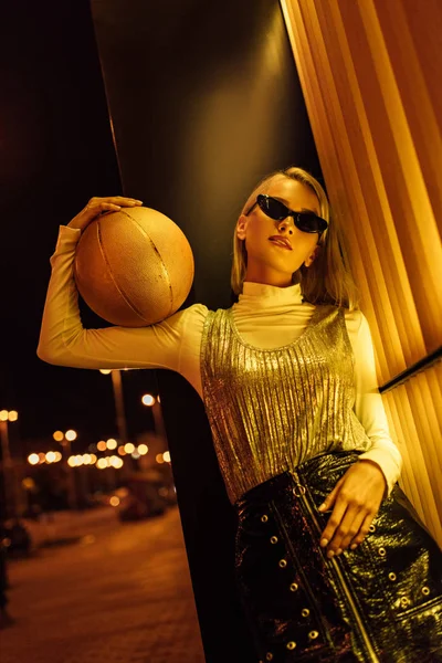 Нижний вид привлекательной женщины в солнечных очках с золотистым глянцевым баскетбольным мячом на улице ночью под желтым светом — стоковое фото