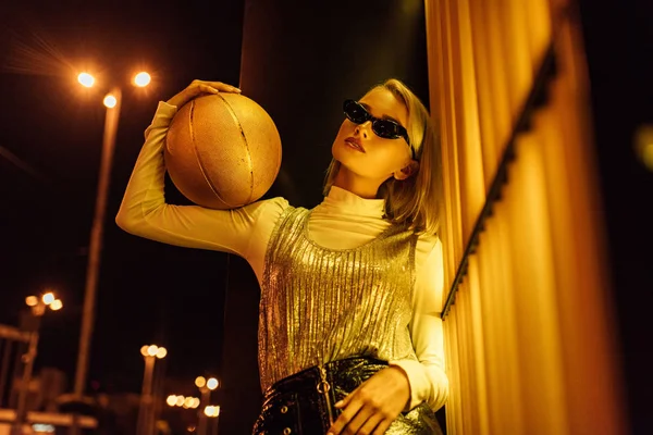 Вид снизу красивой женщины в солнечных очках с золотистым глянцевым баскетбольным мячом на улице ночью под желтым светом — стоковое фото