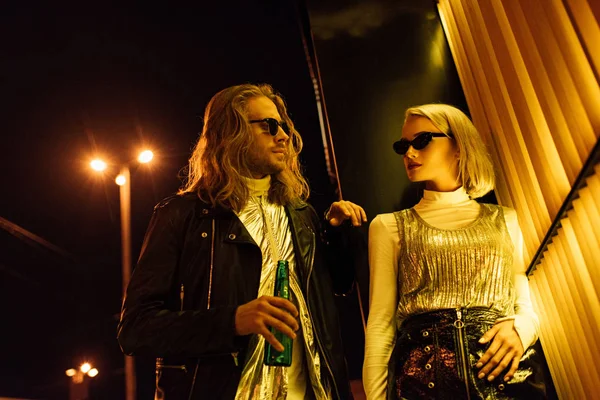 Молодая привлекательная пара в стильной одежде и солнечных очках флиртует на ночной улице под желтым светом — стоковое фото