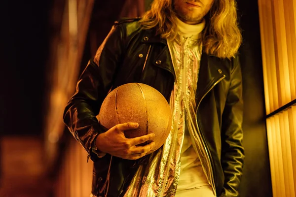 Обрізаний знімок стильного чоловіка в шкіряній куртці, що тримає золотий баскетбольний м'яч під жовтим світлом на вулиці вночі — стокове фото
