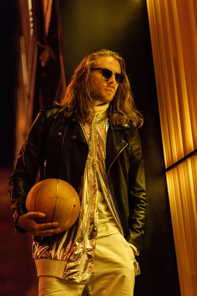 Stylish young man in sunglasses and leather jacket holding golden basketball ball under yellow light on street at night — Stock Photo