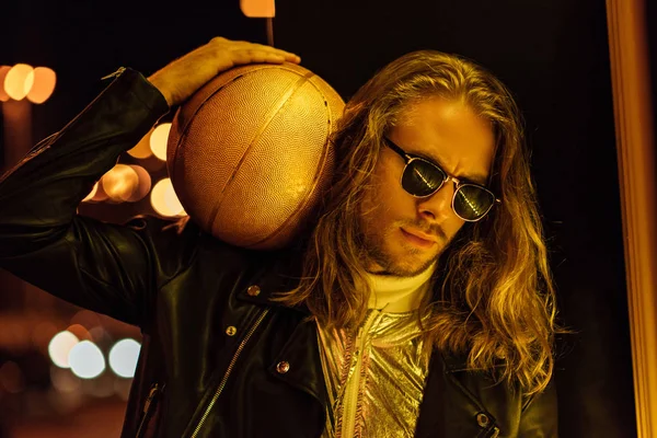 Joven guapo en gafas de sol y chaqueta de cuero que sostiene la bola de baloncesto de oro bajo la luz amarilla en la calle por la noche - foto de stock