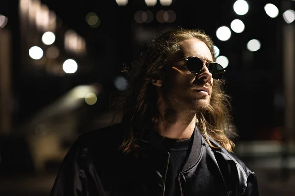 Primer plano retrato de joven atractivo en gafas de sol y chaqueta de cuero en la calle de la ciudad por la noche - foto de stock