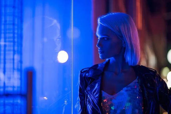 Стильна молода жінка в глянцевому баку зверху і шкіряна куртка на вулиці вночі під блакитним світлом — стокове фото