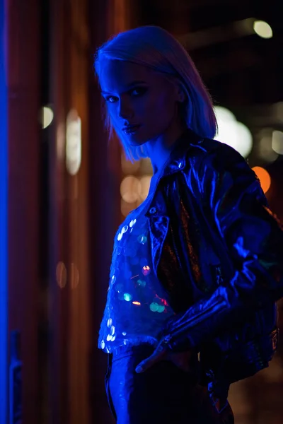Attraktive junge Frau in glänzendem Tank-Top und Lederjacke nachts unter blauem Licht auf der Straße — Stockfoto