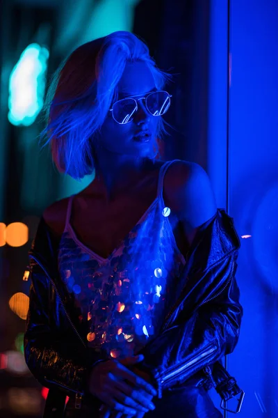 Красива молода жінка в глянцевому баку зверху і сонцезахисні окуляри на вулиці вночі під блакитним світлом — стокове фото