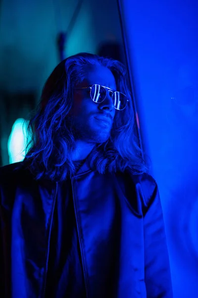 Привлекательный молодой человек в солнечных очках под синим светом на улице ночью — стоковое фото