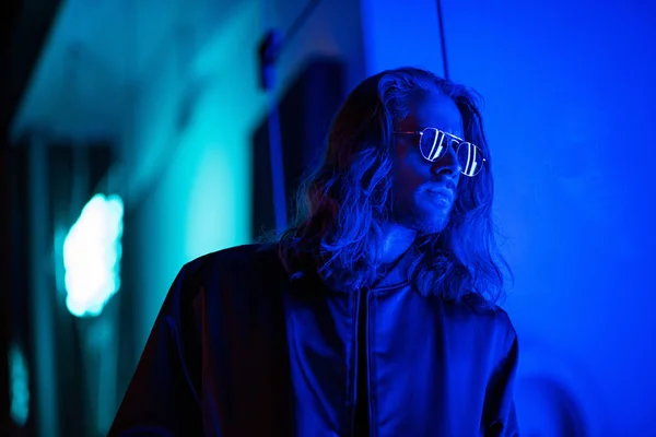 Joven guapo en gafas de sol bajo luz azul en la calle por la noche - foto de stock