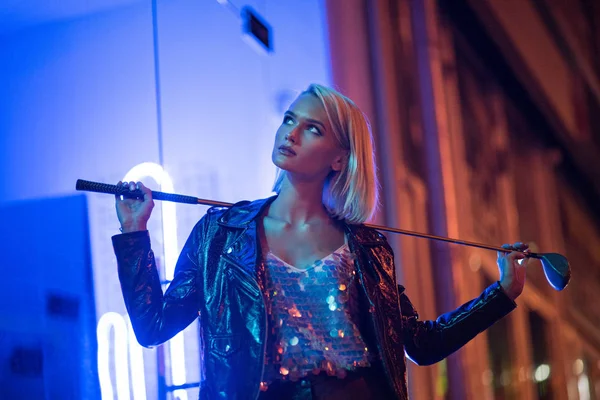 Attraktive junge Frau in Lederjacke, die nachts unter Blaulicht auf der Straße steht und Golfschläger in der Hand hält — Stockfoto
