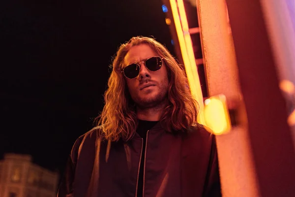 Vista inferior del joven guapo en chaqueta de cuero y gafas de sol en la calle por la noche bajo luz amarilla - foto de stock