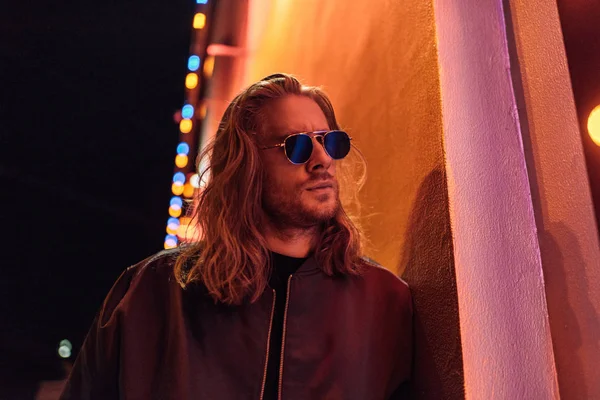 Atractivo joven en chaqueta de cuero y gafas de sol en la calle por la noche bajo luz amarilla - foto de stock