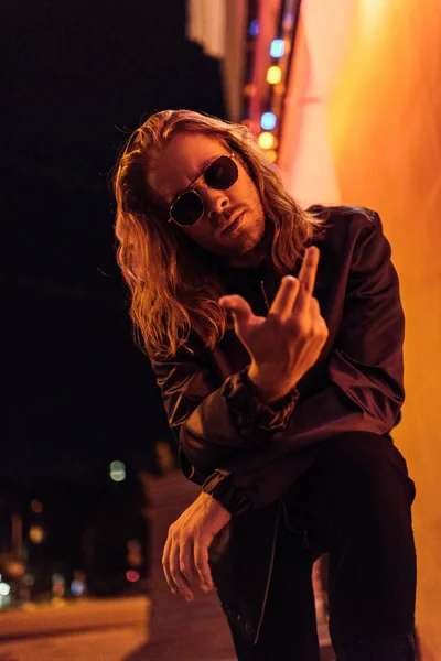 Joven guapo en chaqueta de cuero y gafas de sol que muestran el dedo medio en la cámara en la calle por la noche bajo luz amarilla - foto de stock