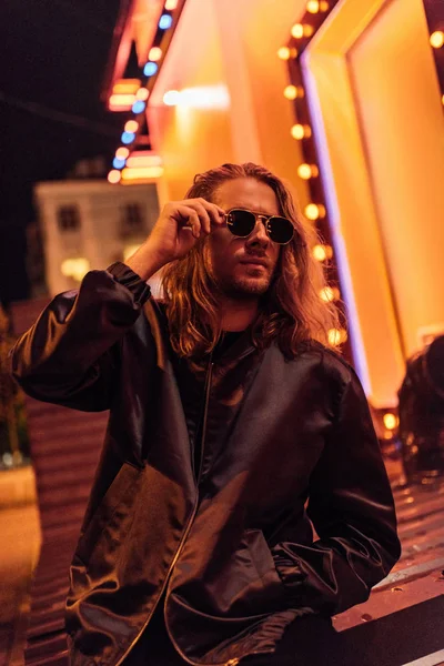 Beau jeune homme en veste en cuir et lunettes de soleil passer du temps seul dans la rue la nuit sous la lumière jaune — Photo de stock