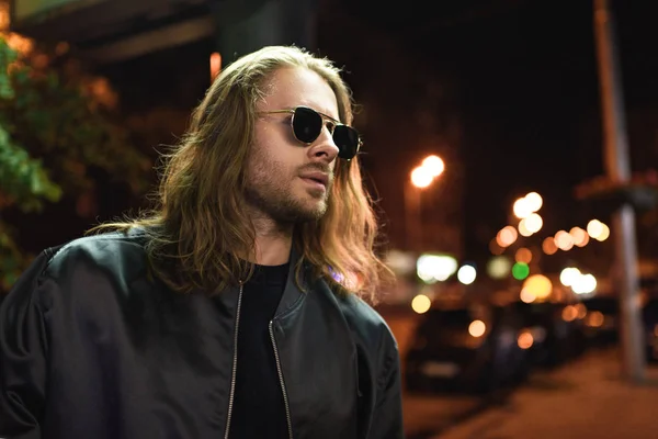 Handsome young man in leather jacket and sunglasses on street at night — Stock Photo