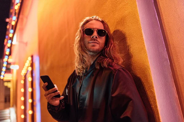 Bottom view of attractive young man in leather jacket and sunglasses using smartphone and looking away on street at night under yellow light — Stock Photo