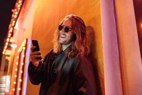 Ridendo giovane in giacca di pelle e occhiali da sole utilizzando smartphone in strada di notte sotto la luce gialla — Foto stock