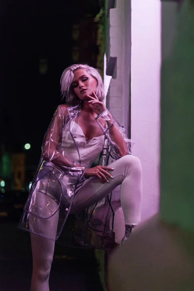 Hermosa mujer joven en impermeable transparente de pie en la calle por la noche bajo la luz rosa - foto de stock