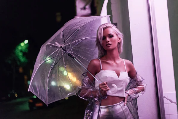 Jovem atraente em capa de chuva transparente com guarda-chuva na rua à noite sob luz rosa — Fotografia de Stock