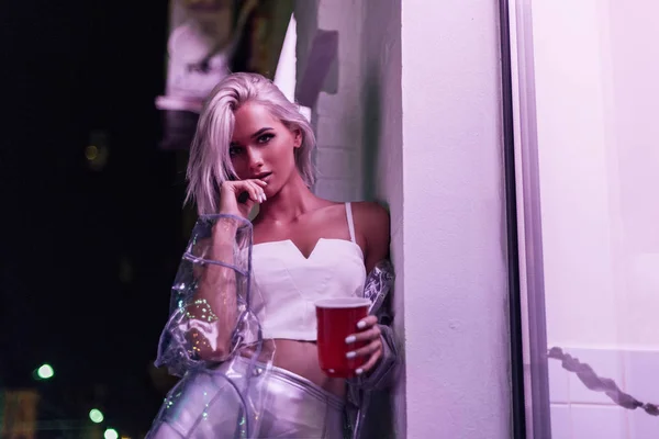 Beautiful young woman with red plastic cup on street at night under pink light — Stock Photo