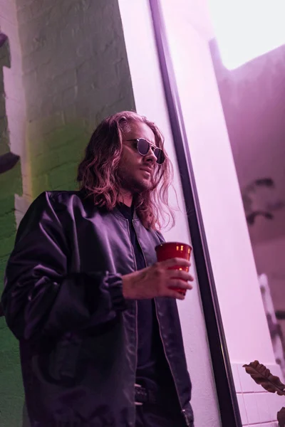 Vista inferior de hombre joven con taza de plástico rojo de bebida en la calle por la noche bajo luz rosa - foto de stock