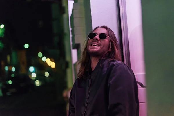 Jeune homme riant en veste en cuir et lunettes de soleil dans la rue la nuit sous la lumière rose — Photo de stock