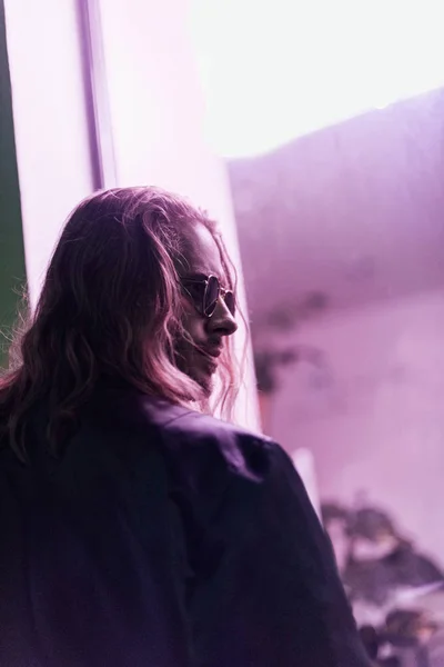 Вид сзади красивого молодого человека в кожаной куртке и солнцезащитных очках, смотрящего ночью на улицу под розовым светом — стоковое фото