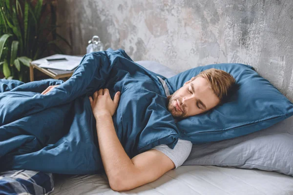Junger schöner Mann schläft unter einer Decke in seinem Bett zu Hause — Stockfoto