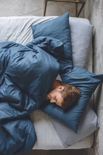 Blick von oben auf einen jungen hübschen Mann, der zu Hause unter einer Decke in seinem Bett schläft — Stockfoto